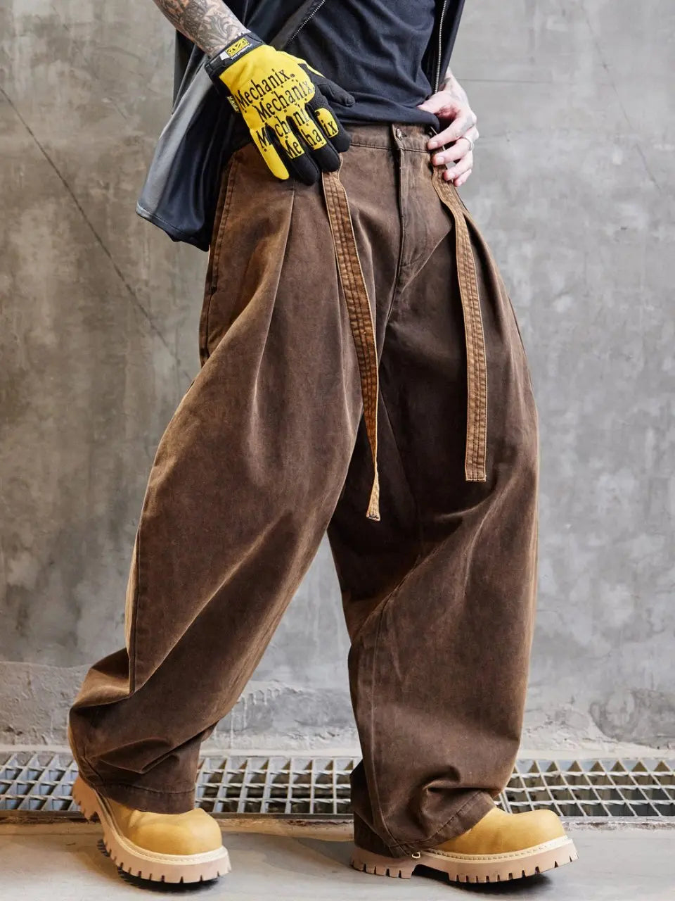 Person wearing brown wide-leg pants and yellow work gloves against a gray concrete wall.