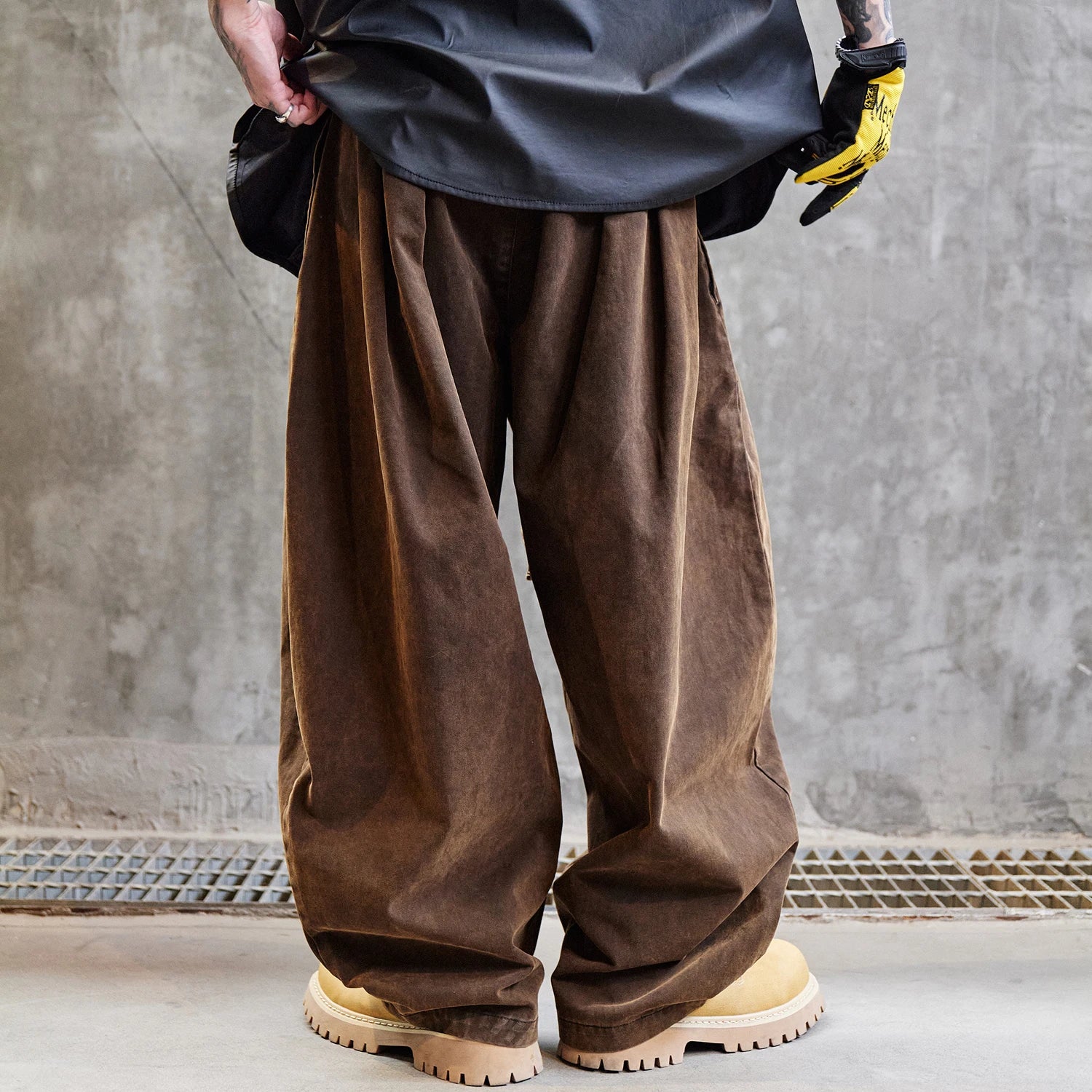 Person wearing brown wide-leg pants and yellow gloves against a gray concrete wall.