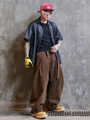 Person wearing a red cap, sunglasses, black shirt, blue jacket, brown pants, and yellow gloves against a gray concrete wall.