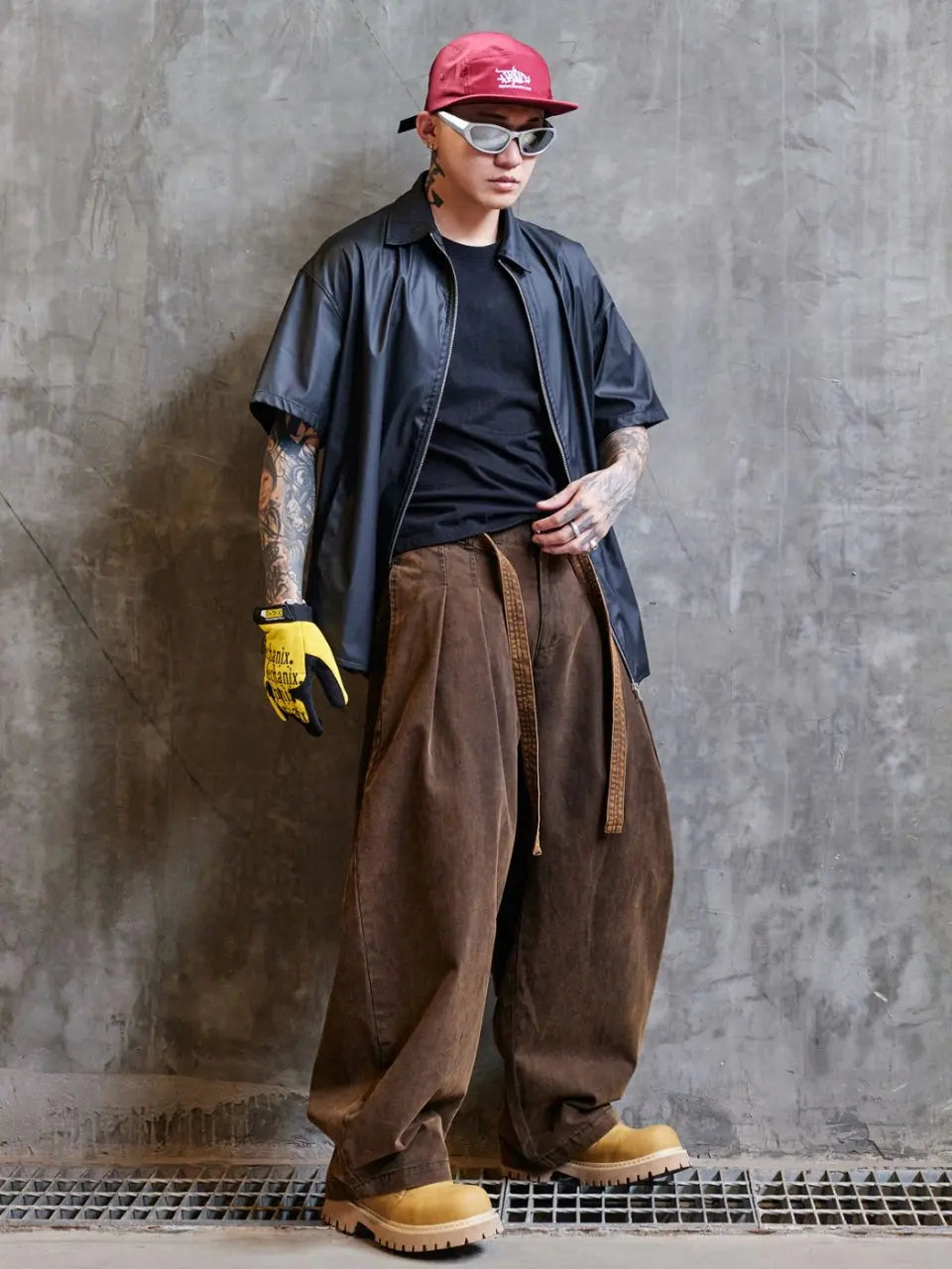 Person wearing a red cap, sunglasses, black shirt, blue jacket, brown pants, and yellow gloves against a gray concrete wall.