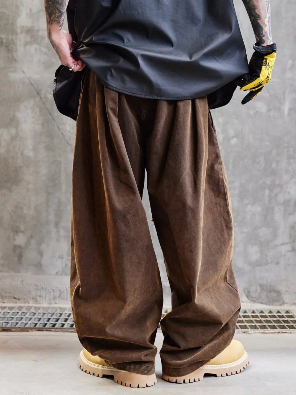 Person wearing brown wide-leg pants and a dark jacket against a gray concrete wall.