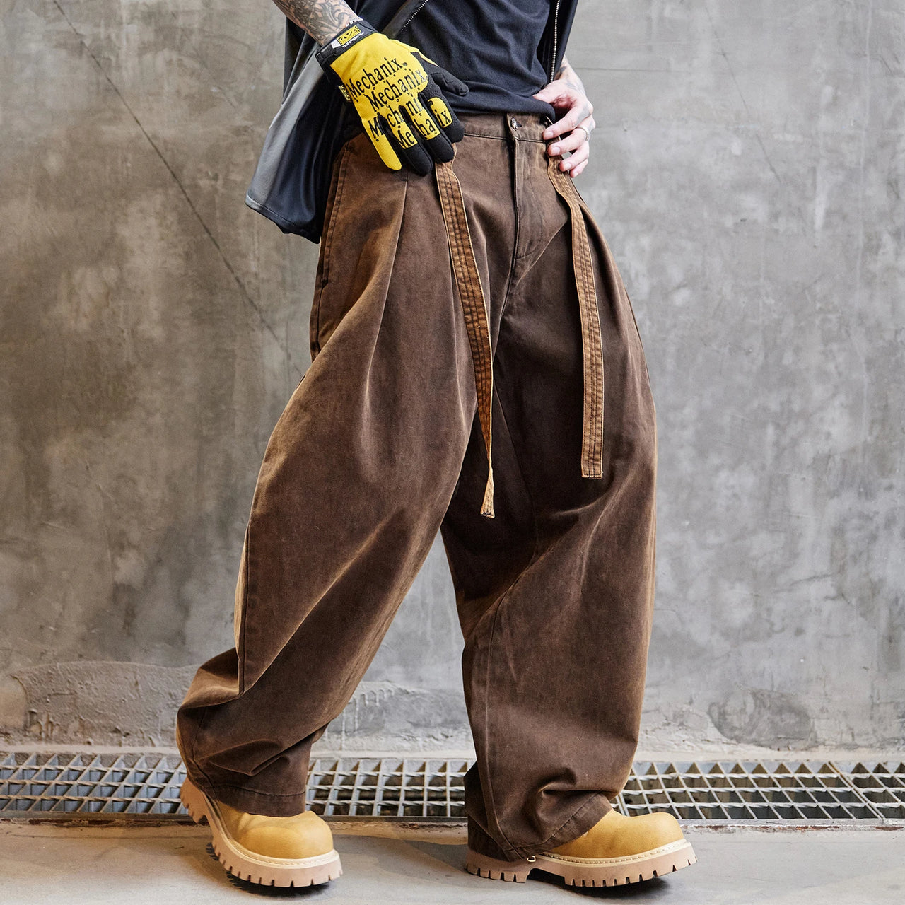Person wearing brown baggy pants and yellow work gloves against a gray concrete wall.