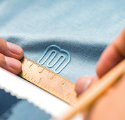 Close-up of a hand holding a ruler against blue fabric with a brand logo.