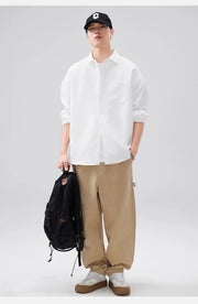 Person wearing a white shirt, beige pants, and a black cap, holding a black backpack on a light gray background