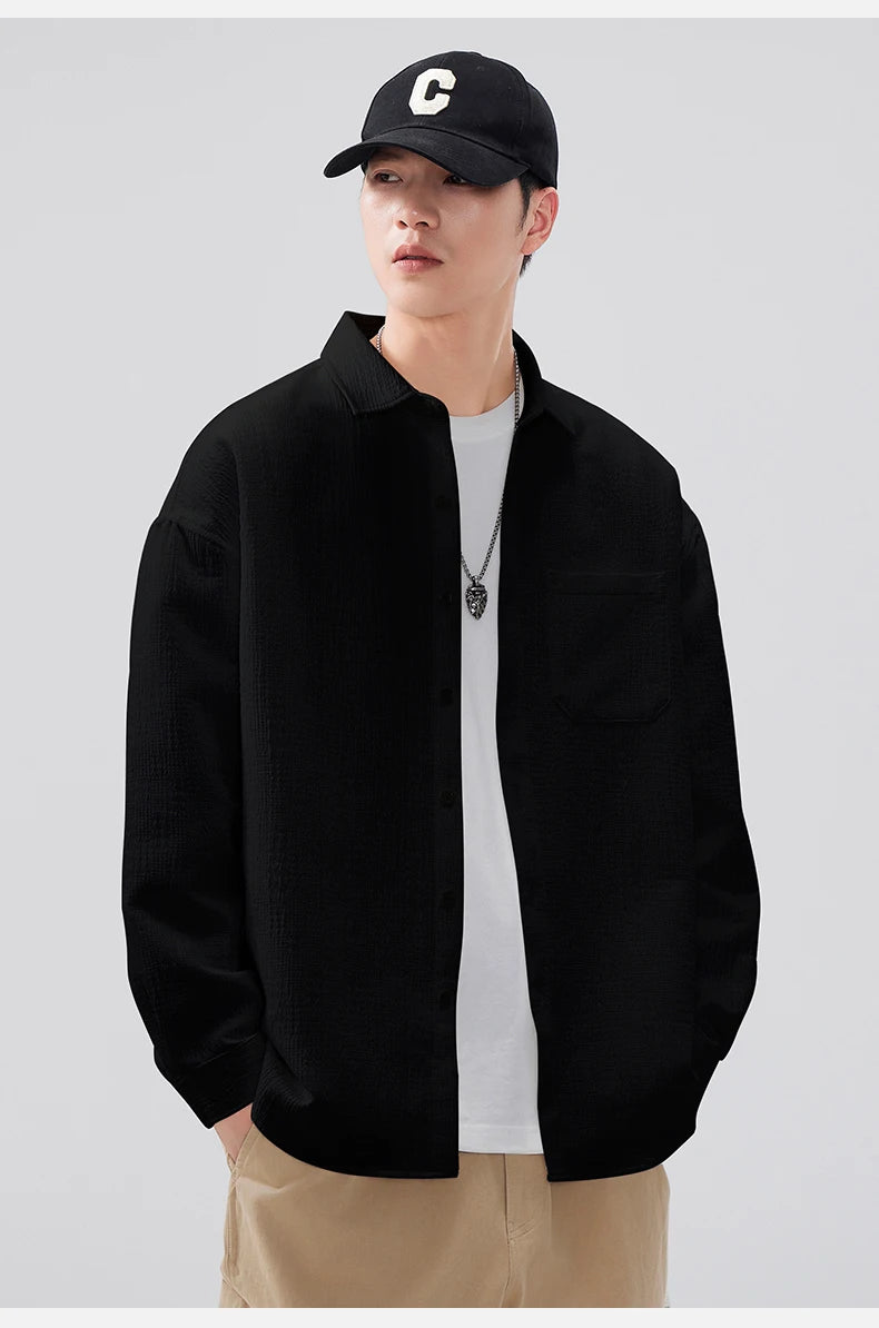 Person wearing a black jacket, white shirt, beige pants, and a black cap with a white logo on a light gray background