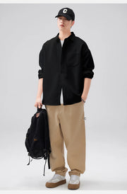 Person wearing a black shirt, beige pants, and a black cap, holding a black backpack on a light gray background