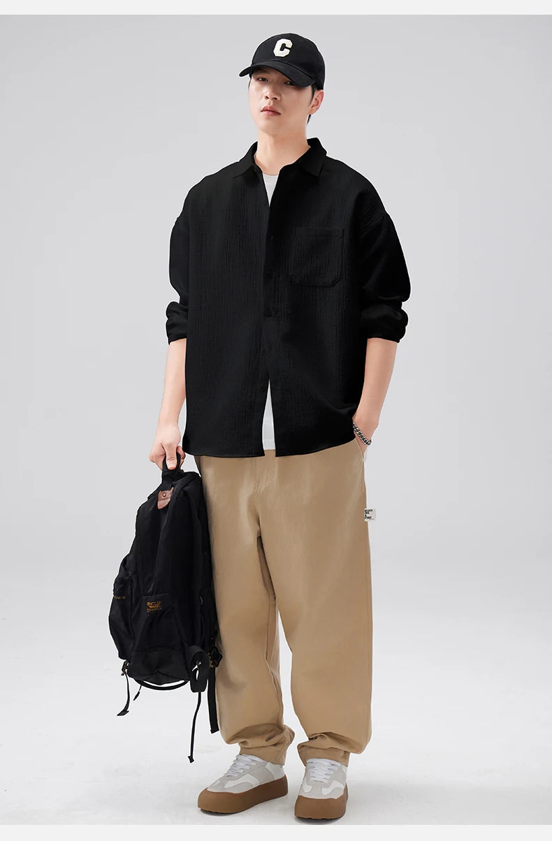 Person wearing a black shirt, beige pants, and a black cap, holding a black backpack on a light gray background