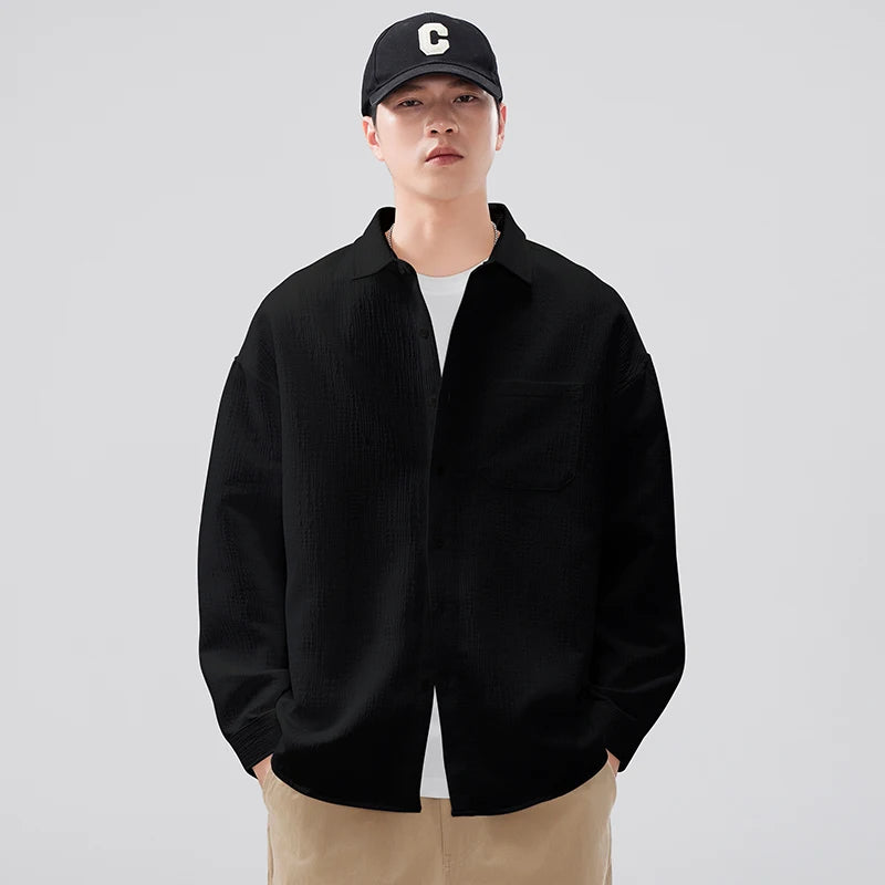 Person wearing a black jacket, white shirt, and beige pants with a black cap featuring a white logo on a light gray background.