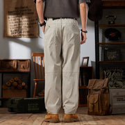 Person wearing beige pants standing in a room with wooden furniture and a backpack.