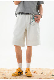 A person wearing light beige rolled-calf shorts with a decorative patterned belt, a gray t-shirt, white socks, and yellow sneakers, standing on a sandy surface against a white background.
