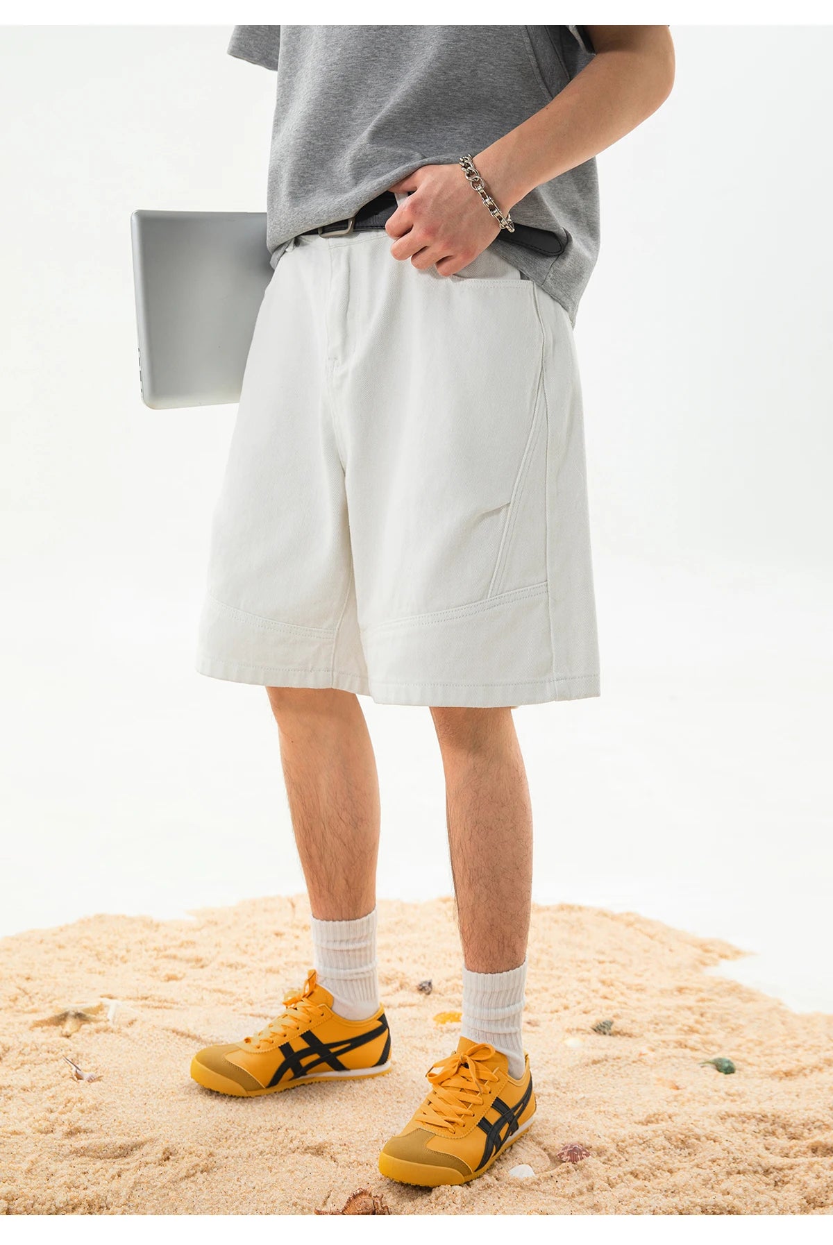 A person wearing light beige or off-white casual shorts, a gray t-shirt, white socks, and yellow sneakers with black stripes, standing on sand against a white studio background.
