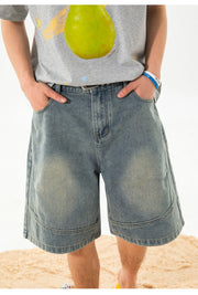 A person wearing light-wash blue denim shorts with a gray graphic t-shirt, standing on a sandy beach against a white background.