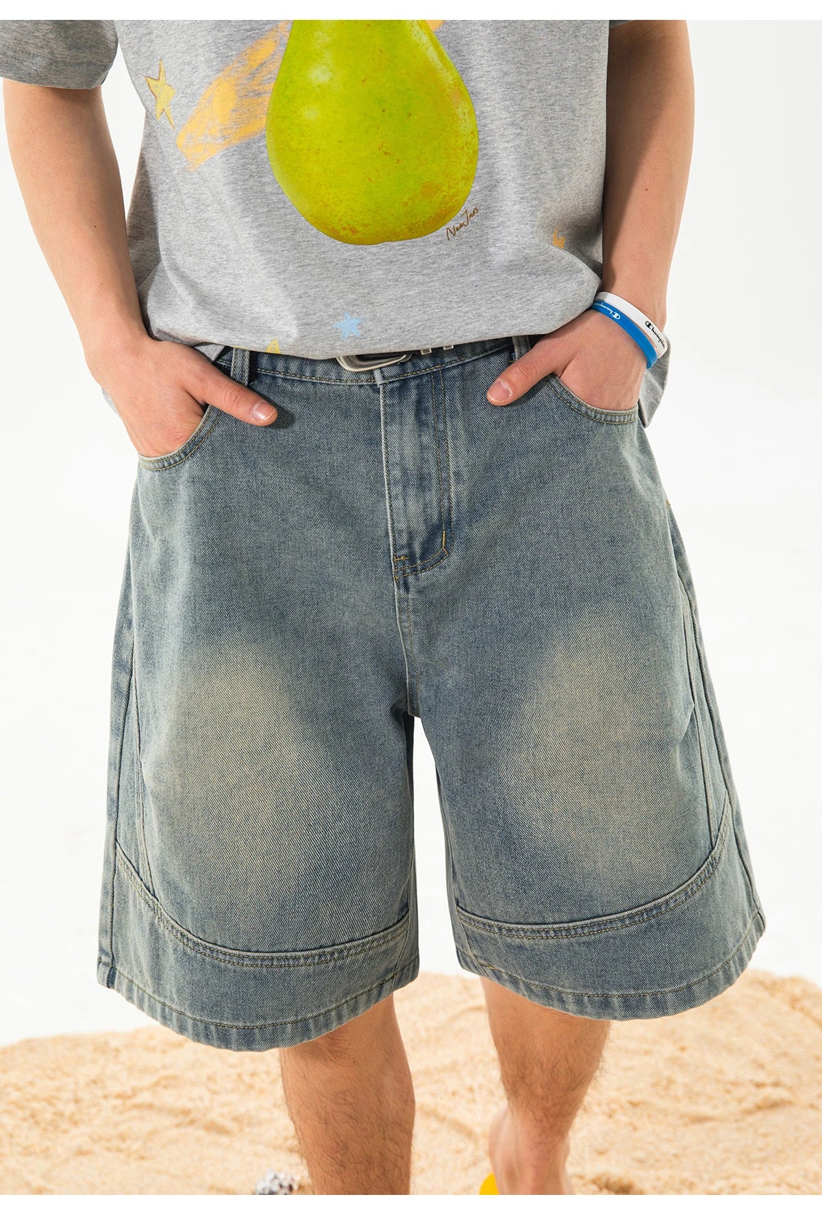A person wearing light-wash blue denim shorts with a gray graphic t-shirt, standing on a sandy beach against a white background.