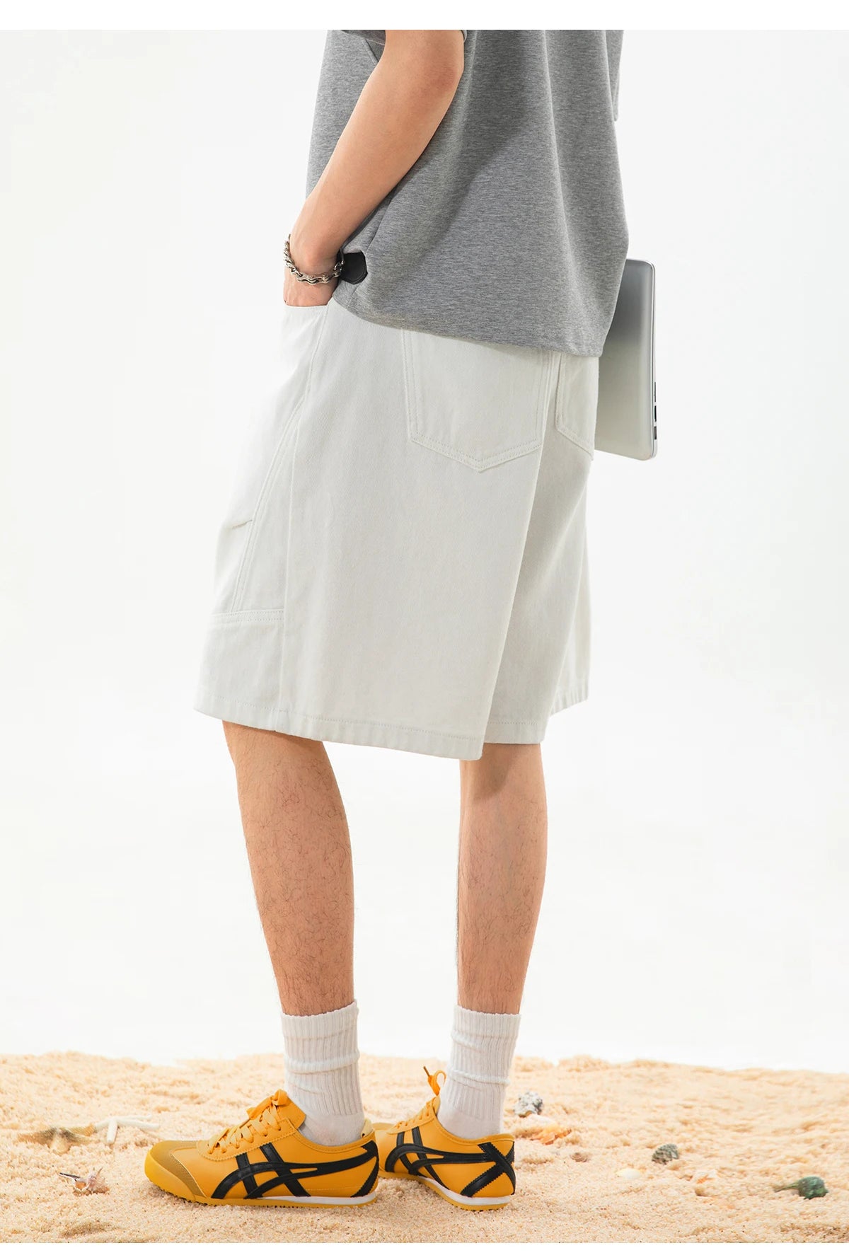 A person standing on sand wearing light gray knee-length shorts, a heather gray sleeveless top, white socks, and yellow athletic shoes with black stripes.