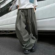 A person wearing very loose, oversized dark olive green cargo-style pants with a white t-shirt and black shoes, standing on asphalt in front of a white vehicle.