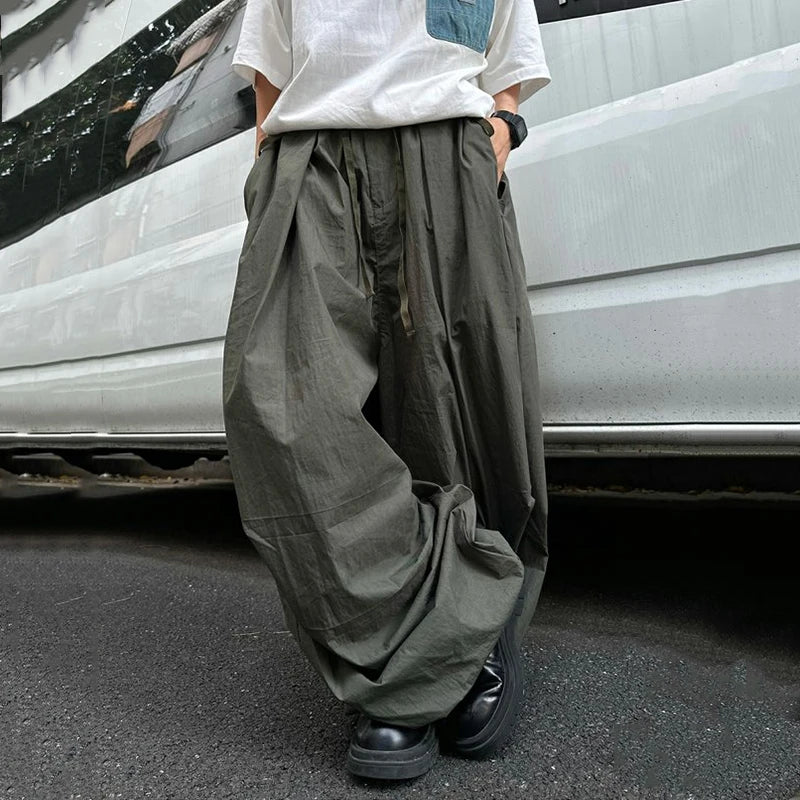 A person wearing very loose, oversized dark olive green cargo-style pants with a white t-shirt and black shoes, standing on asphalt in front of a white vehicle.