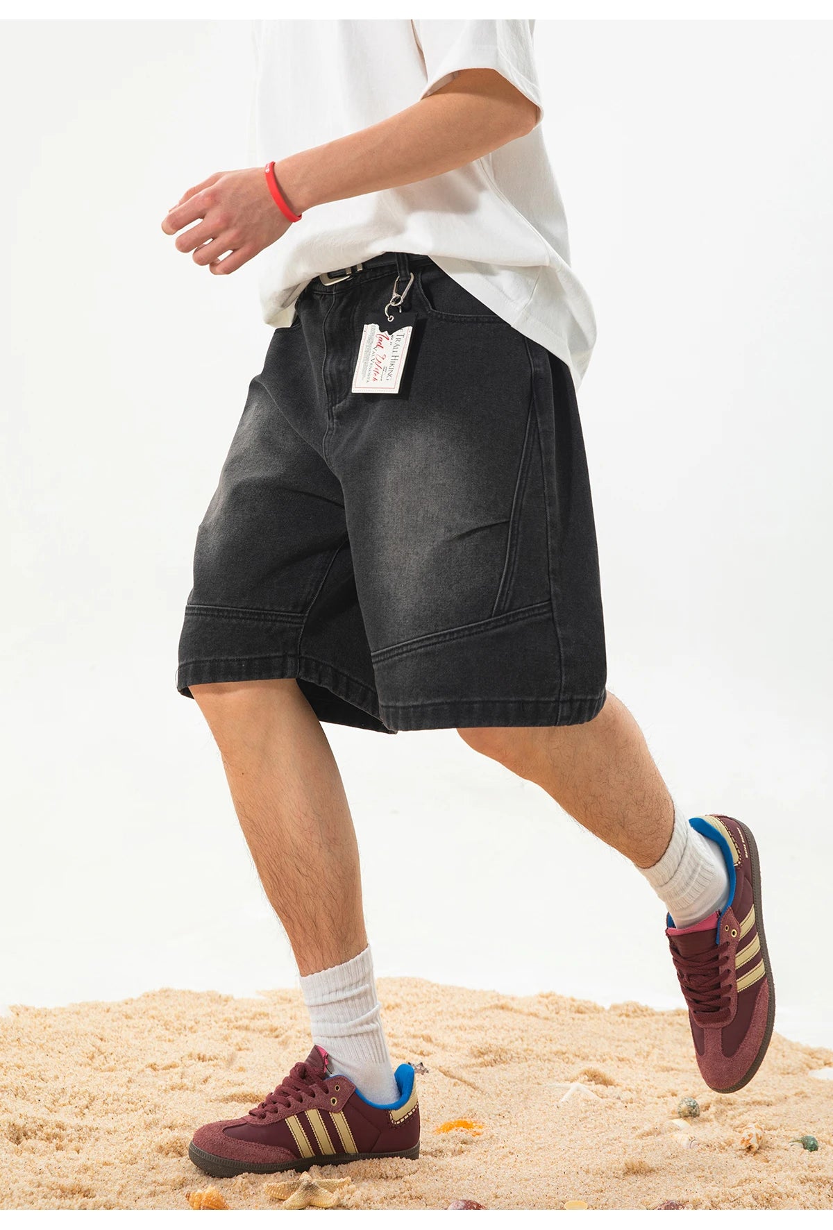 A person wearing black denim shorts, a white t-shirt, white socks, and maroon sneakers with gold stripes, standing on sand against a white background.