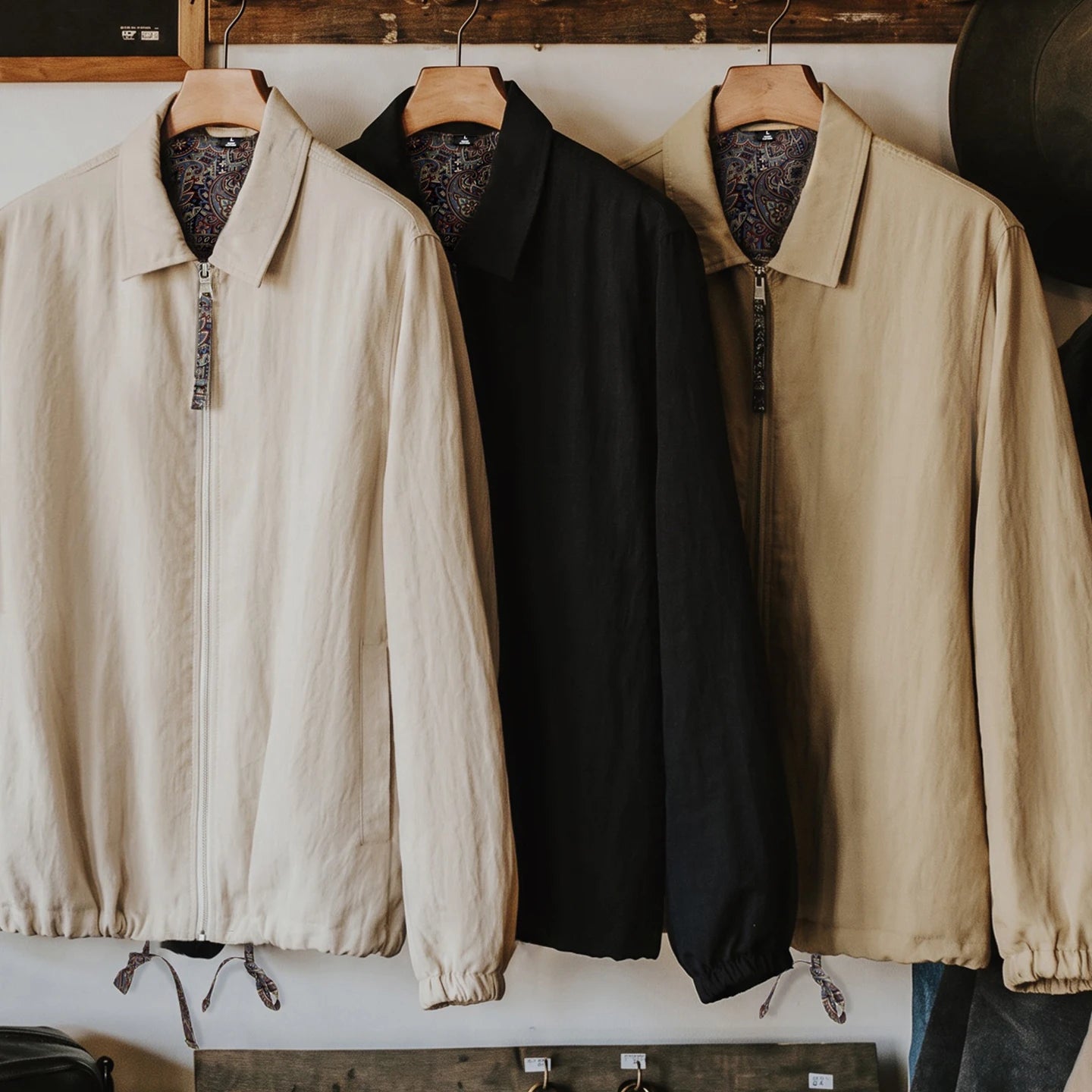 Three lightweight zip-front jackets hanging on wooden hangers against a white wall. The left jacket is light beige, the center jacket is black, and the right jacket is tan.