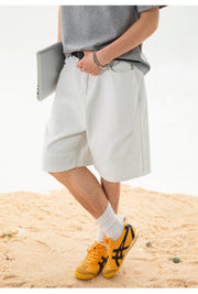 A person standing on sand wearing light gray shorts, a gray t-shirt, white socks, and yellow sneakers, holding a silver laptop.