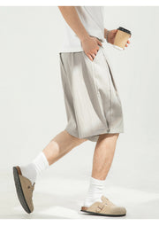 A person wearing light beige pleated linen shorts, white crew socks, and beige slip-on clogs, holding a paper coffee cup against a clean white background.