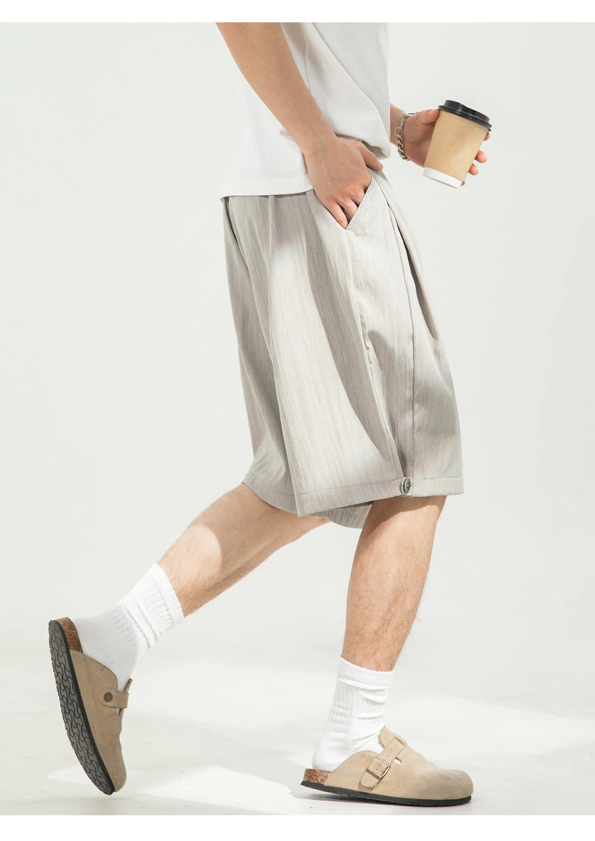 A person wearing light beige pleated linen shorts, white crew socks, and beige slip-on clogs, holding a paper coffee cup against a clean white background.