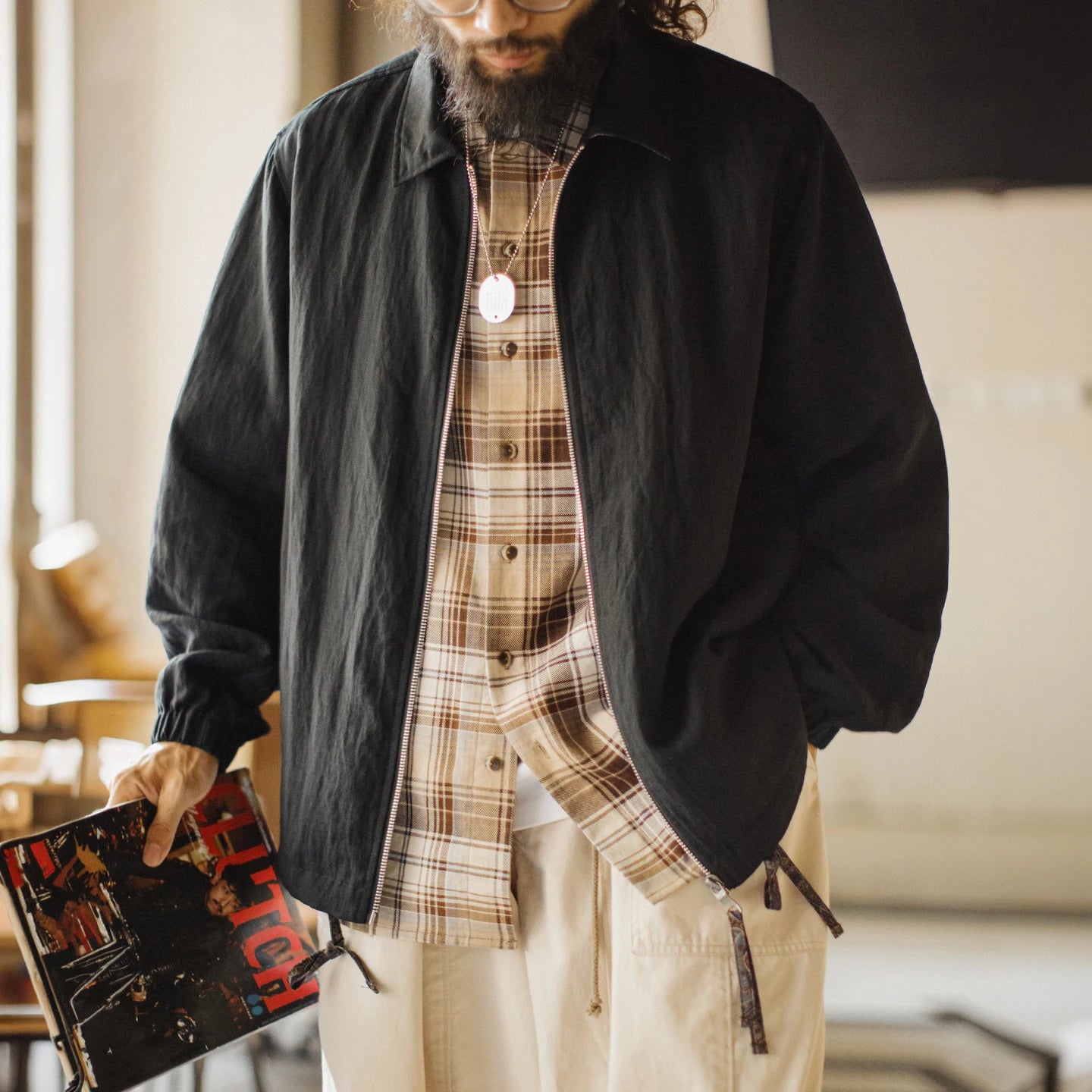 A man wearing a black lightweight overshirt over a brown and beige plaid button-up shirt, paired with cream cargo pants, holding a book in a softly lit indoor setting.