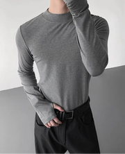 Person wearing a gray long-sleeve shirt and dark pants against a neutral background