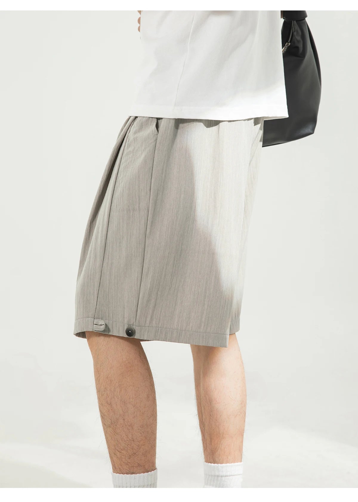 A person wearing light gray pleated shorts with a white top and white socks, holding a black shoulder bag against a plain light background.