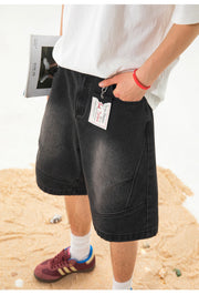 A person wearing faded black denim shorts with a white t-shirt and maroon sneakers, standing on a sandy beach. The shorts feature a visible hangtag and a pocket with a red wristband nearby.