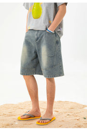 A person wearing light-wash blue denim shorts with a gray graphic t-shirt and yellow flip-flops, standing on a sandy surface against a white background.