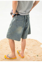 A person standing on sand wearing faded blue denim shorts, a gray t-shirt, and yellow flip-flops, shown from the back.