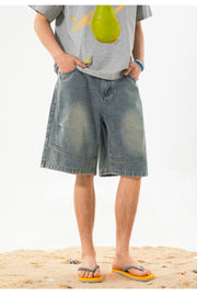A person wearing light-wash blue denim shorts with faded yellowish patches, a gray graphic t-shirt, and yellow flip-flops standing on a sandy surface against a white background.