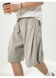 A person wearing light gray pleated knee-length shorts with an elastic waistband, paired with a white t-shirt and holding a coffee cup, against a plain white background.
