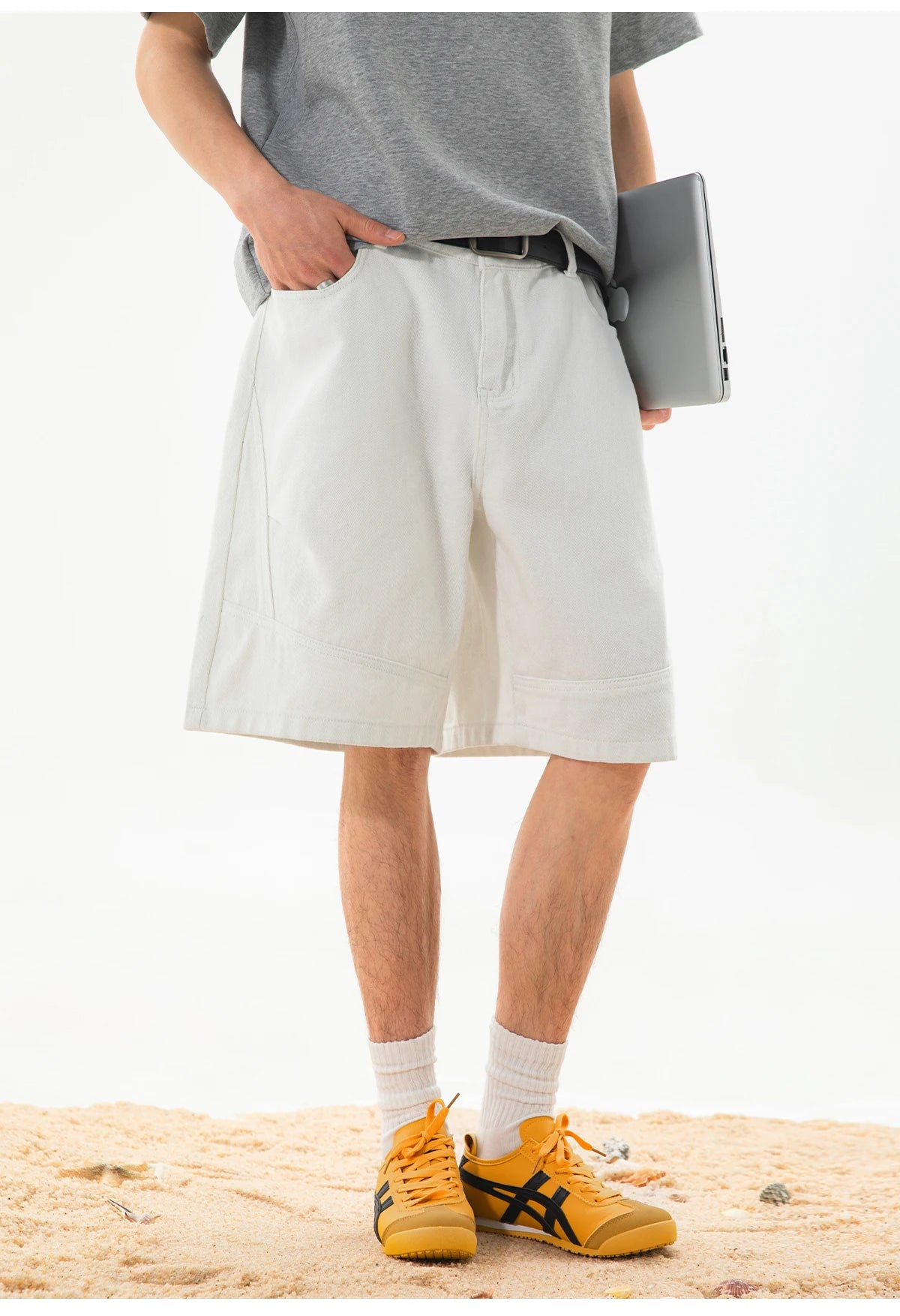 A person standing on sand wearing light beige wide-leg shorts, a gray t-shirt, white socks, and yellow sneakers, holding a gray laptop.