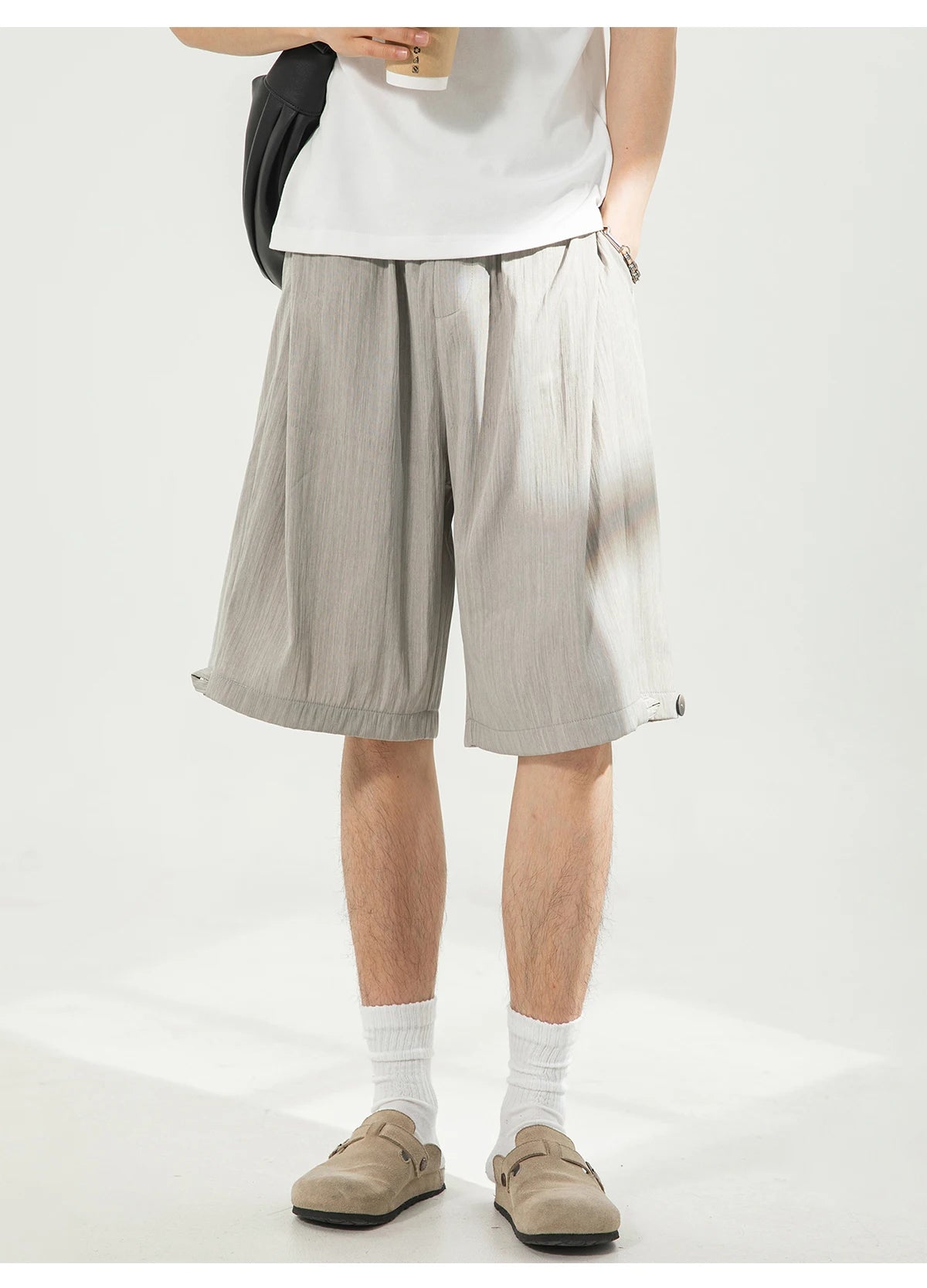 A person wearing light gray pleated linen shorts, styled with a white t-shirt, white socks, and beige loafers, standing against a white studio background.