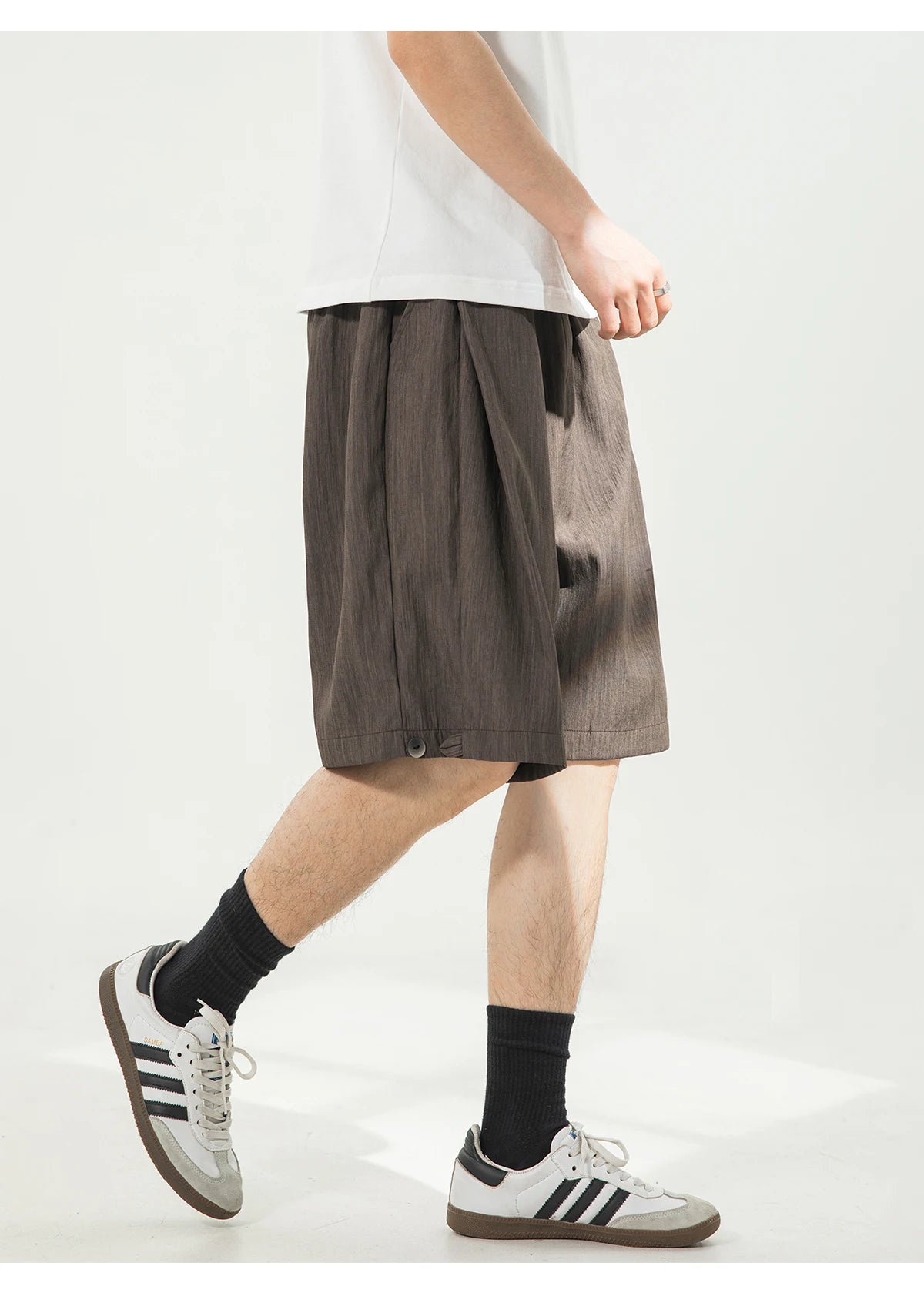 A person wearing loose, pleated brown shorts with a white t-shirt, black socks, and white Adidas sneakers, shown from the waist down against a light gray studio background.