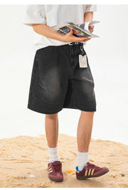A person standing on sand wearing black denim shorts, a white t-shirt, white socks, and burgundy sneakers with gold stripes, holding an open magazine.