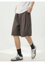 A person wearing loose dark brown pleated shorts, a white t-shirt, black socks, and white sneakers with black stripes, standing against a light gray studio background.