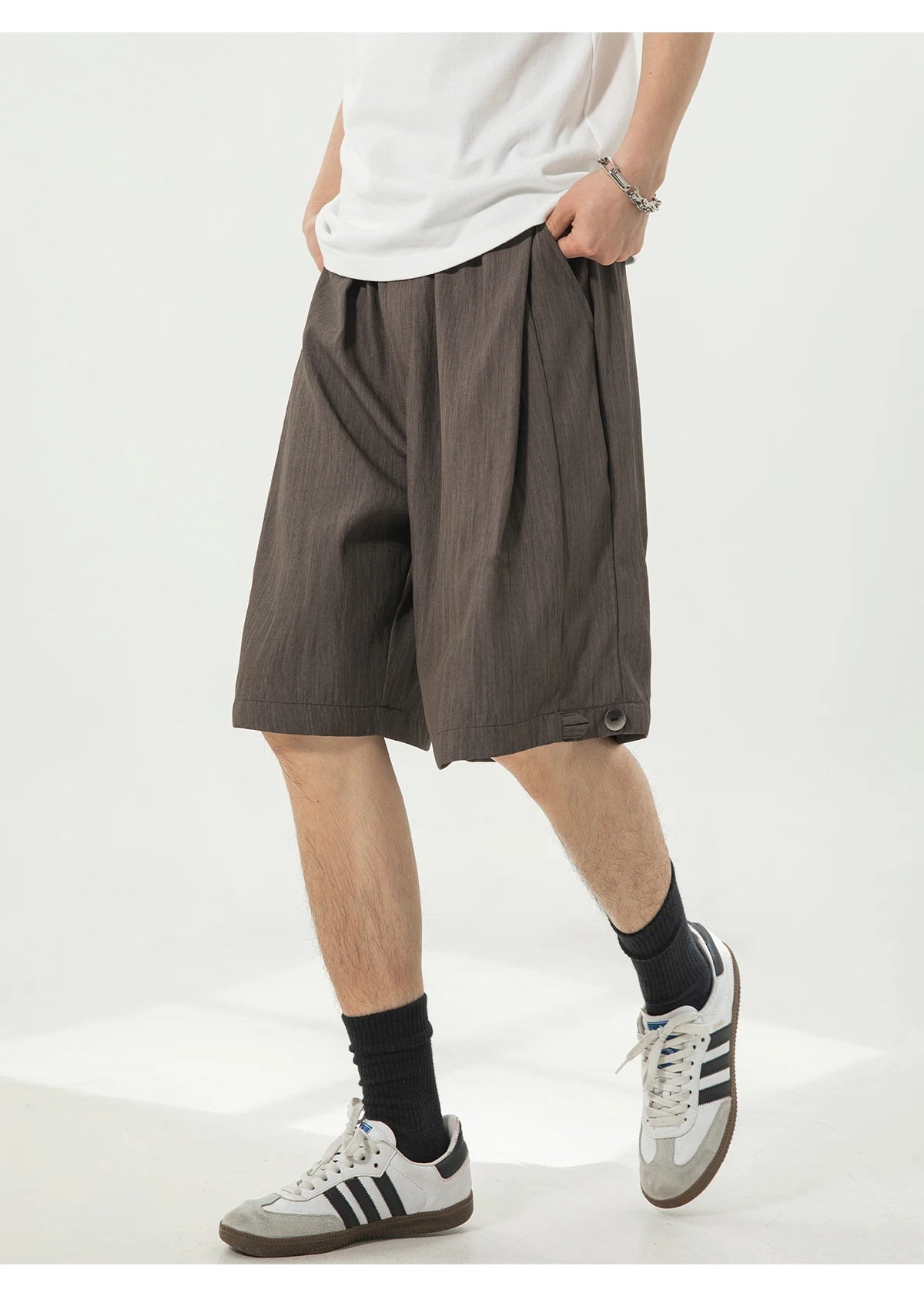 A person wearing loose dark brown pleated shorts, a white t-shirt, black socks, and white sneakers with black stripes, standing against a light gray studio background.