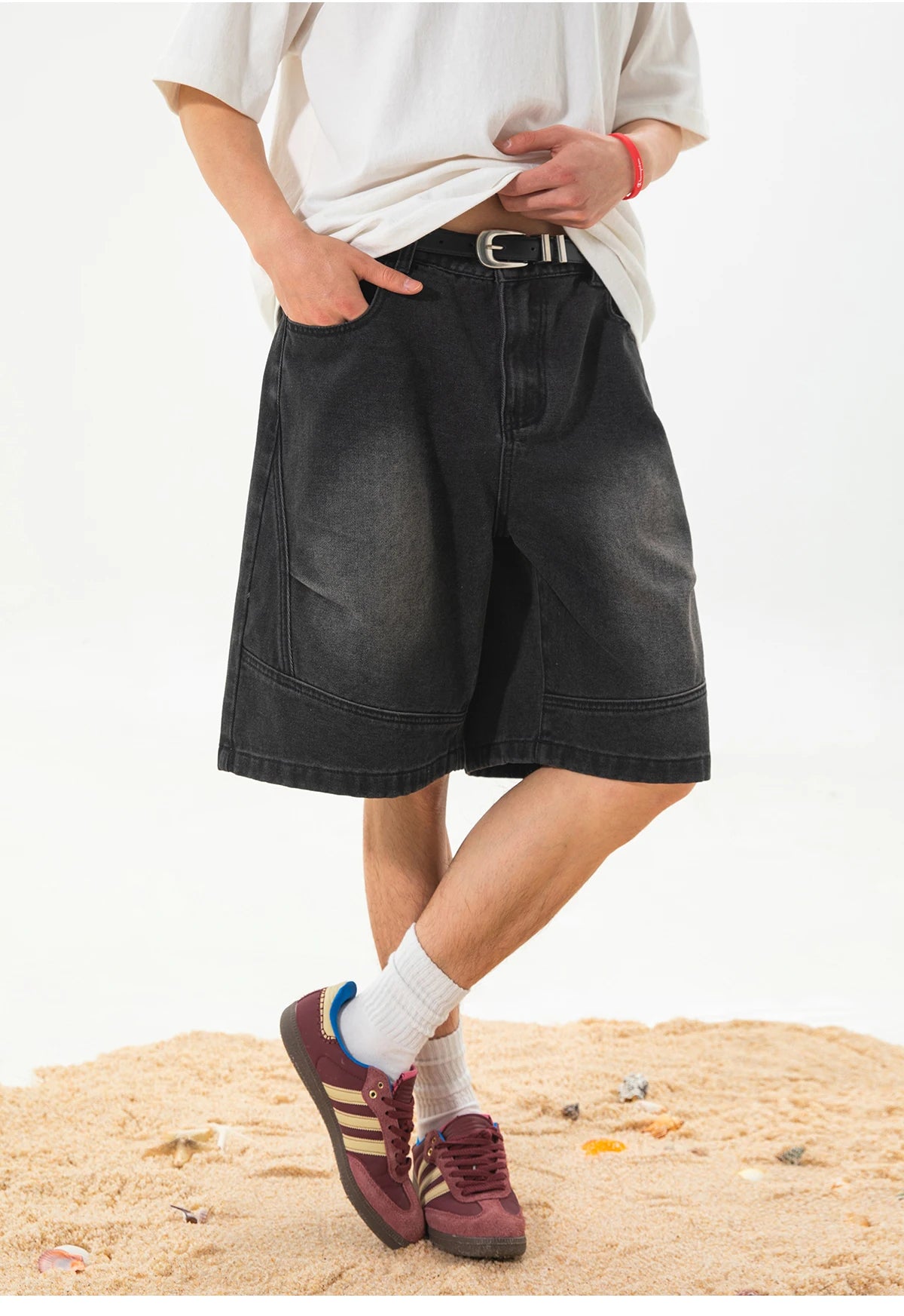 A person wearing oversized black denim shorts with a belt, a white t-shirt, white socks, and burgundy sneakers, standing on a sandy surface against a plain white background.