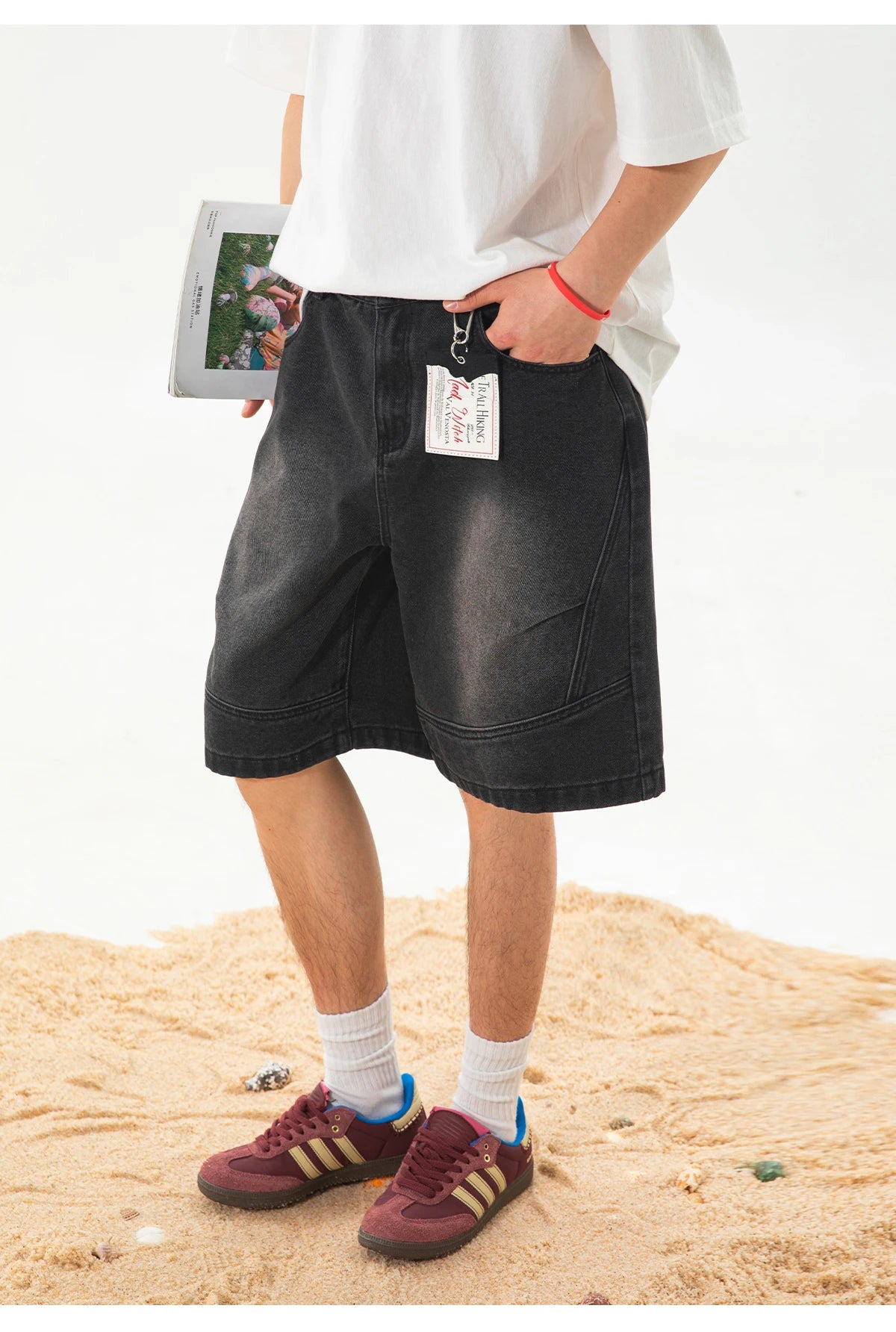 A person wearing black denim shorts, a white t-shirt, white socks, and burgundy sneakers with gold stripes, standing on a sandy surface against a white background.