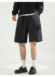 A person wearing black pleated shorts, a black sweatshirt, white socks, and black leather shoes, shown from the waist down against a light gray studio background.