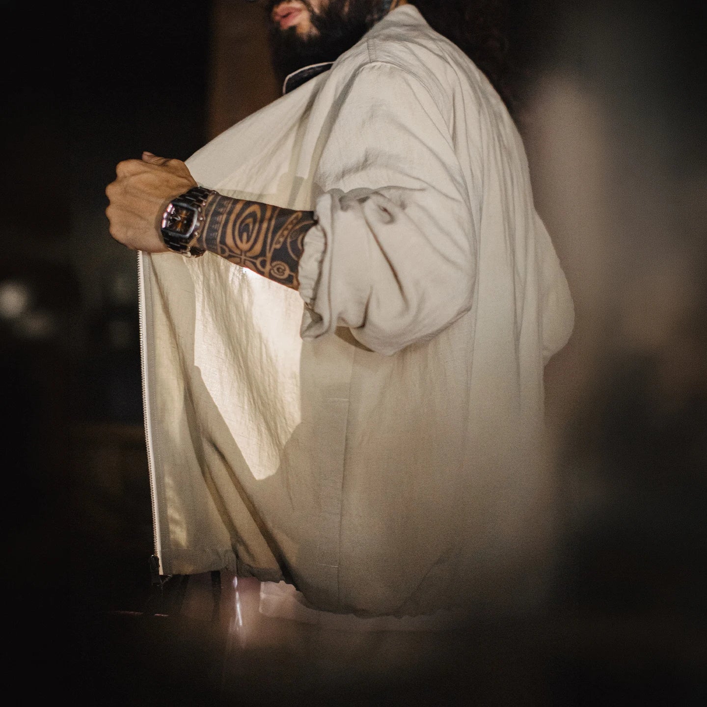 A man wearing a light beige linen shirt with a black watch on his tattooed forearm, shown in a moody, low-light setting.