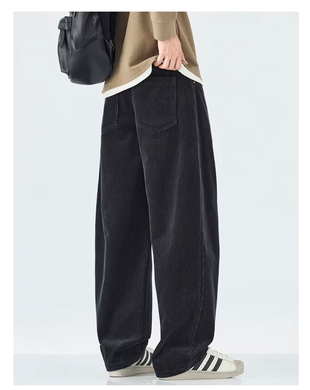 A person wearing loose black corduroy pants, styled with a beige top, white sneakers, and a black backpack, shown from the waist down against a light gray studio background.