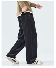 A person wearing loose black corduroy pants, a beige long-sleeve top, and white sneakers with black stripes, shown from the waist down against a light gray studio background.