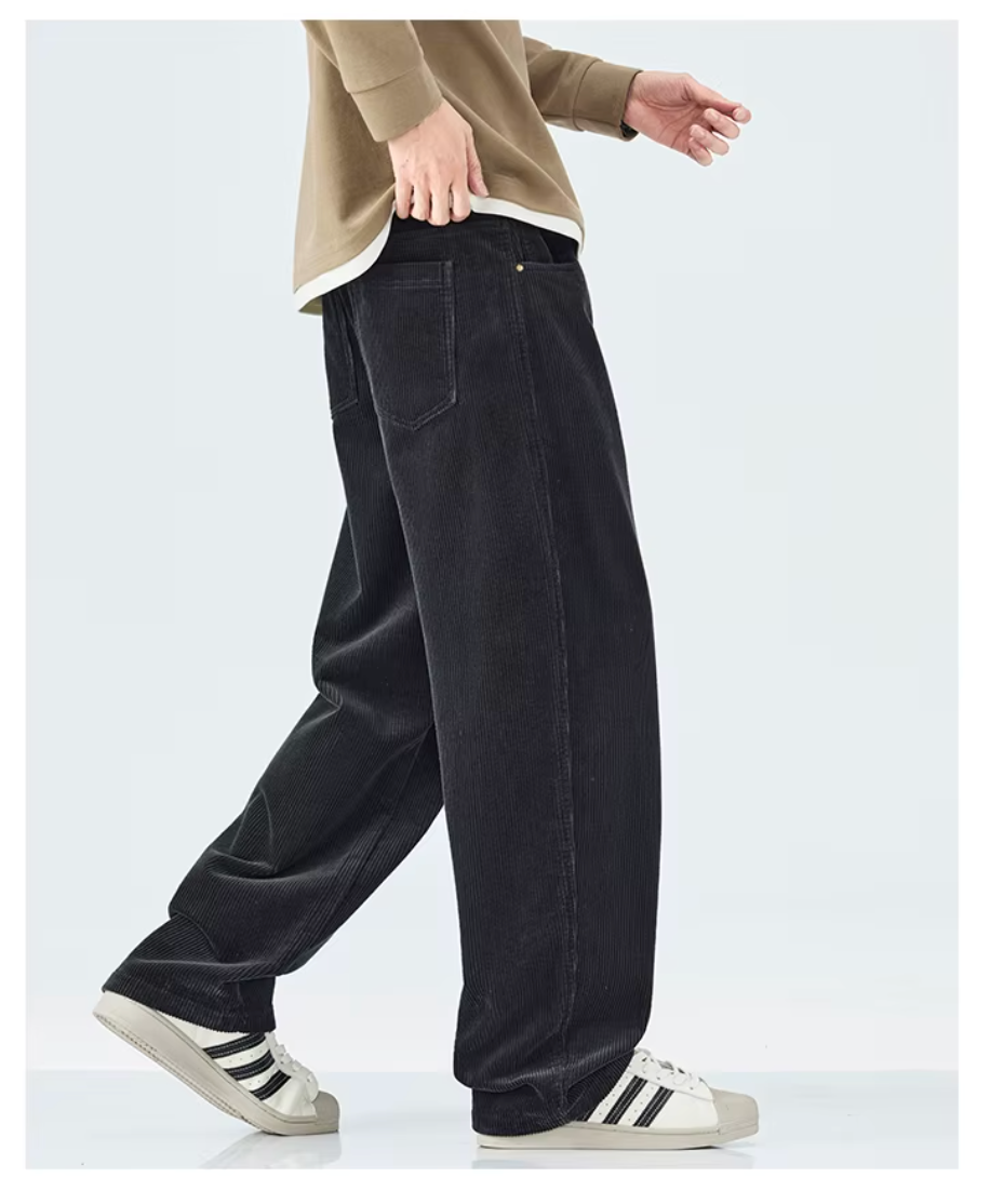 A person wearing loose black corduroy pants, a beige long-sleeve top, and white sneakers with black stripes, shown from the waist down against a light gray studio background.