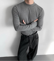 Person wearing a gray long-sleeve shirt and black pants against a white wall.