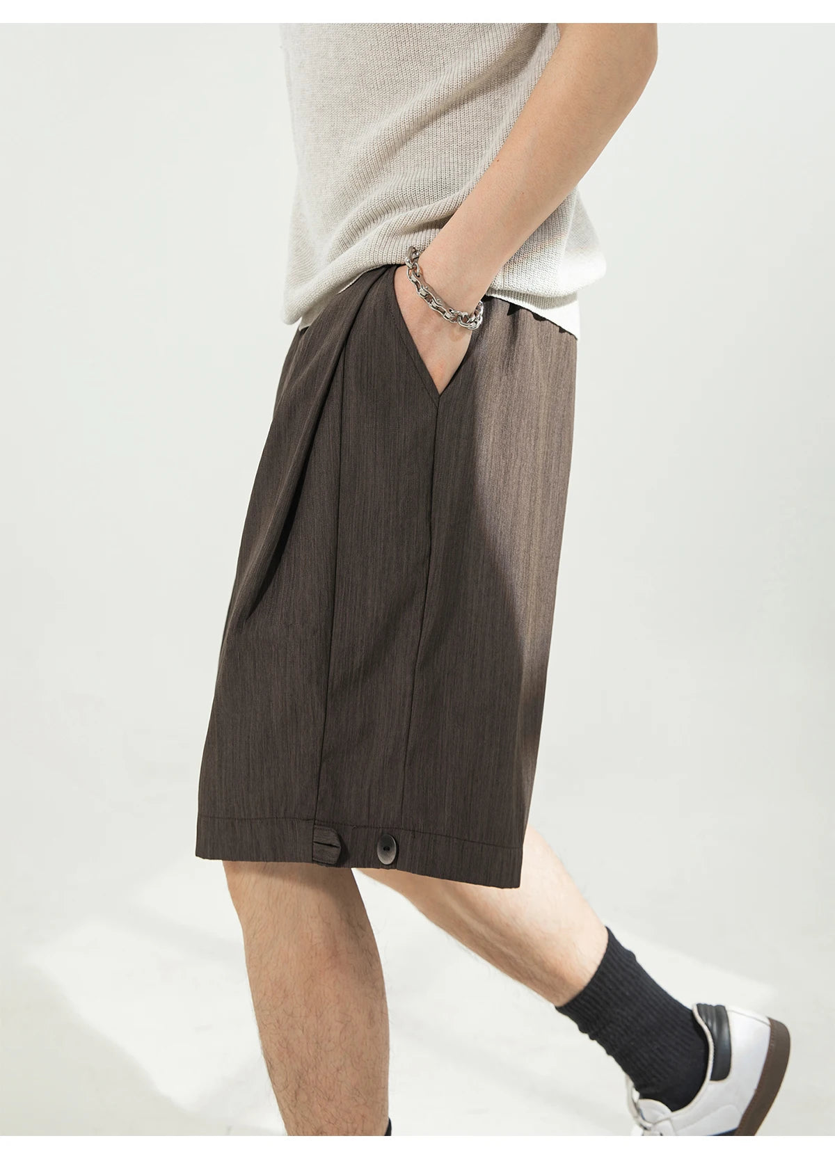 A person wearing dark brown pleated shorts with a split front, side pockets, and button details at the hem, styled with a light gray knit top, black socks, and white sneakers against a plain light background.