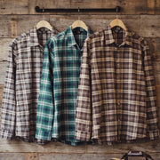 Three long-sleeve plaid button-up shirts hanging on wooden hangers against a rustic wood plank wall. The left shirt is taupe and gray, the middle is green and blue, and the right is brown and beige.