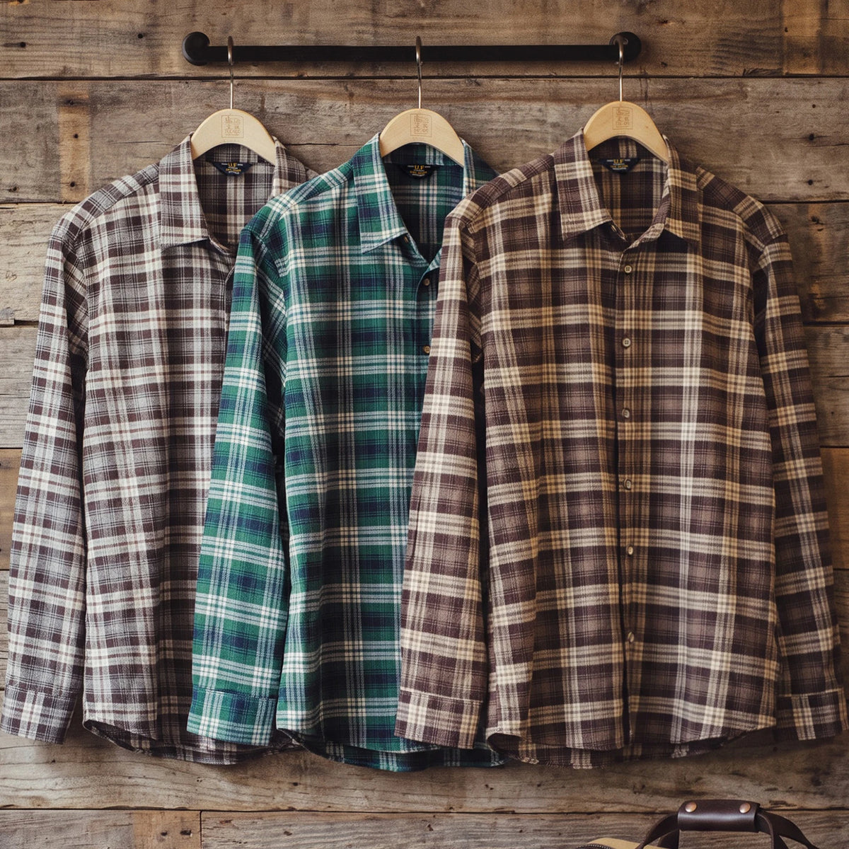 Three long-sleeve plaid button-up shirts hanging on wooden hangers against a rustic wood plank wall. The left shirt is taupe and gray, the middle is green and blue, and the right is brown and beige.